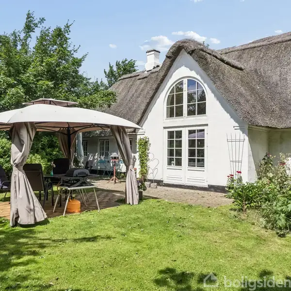 Et hvidt stråtækt hus står i en have med grøn plæne. Foran huset ses en pavillon med borde og stole, omgivet af buske og træer under en klar himmel.