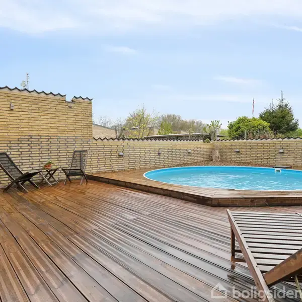 Rund pool omgivet af en træterrasse med liggestole og et bord. Omkranset af en gule murstensvægge i en baghave med grønne planter i baggrunden.