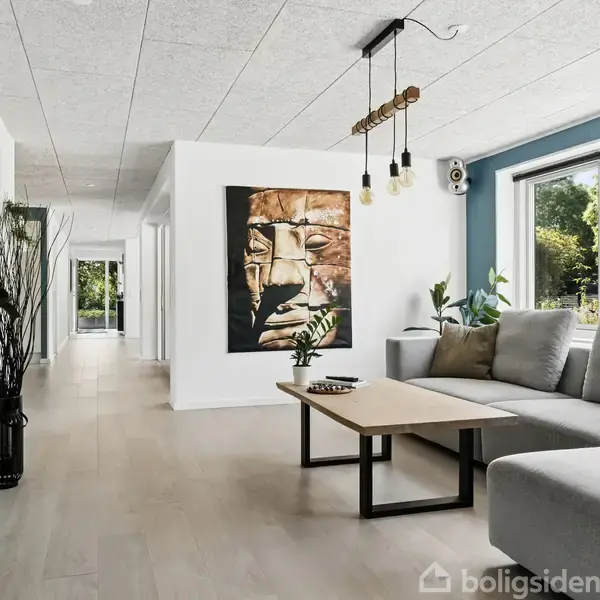 Grå hjørnesofa placeret ved siden af stort vindue. Foran sofaen står et sofabord. Maleri af ansigt hænger på væggen. Stue dekoreret med planter og moderne lamper.