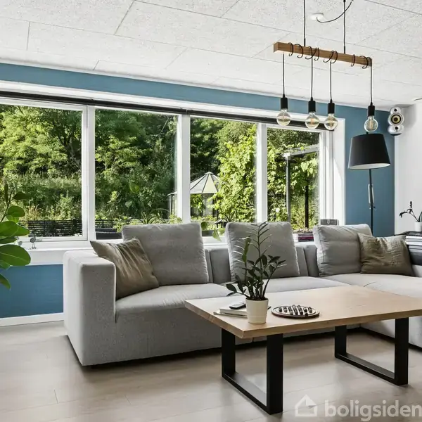 Sofa i lysegrå står foran store vinduer med udsigt til grøn have. Stue med træbord, loftlamper og plante. Højttalere på væggen, skrivebord med kort på væggen.