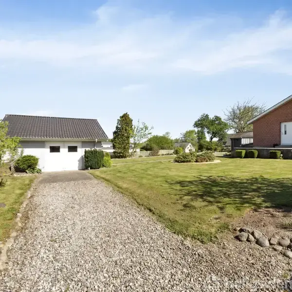 Grusvej fører til garage ved en hvid bygning med sort tag. Til højre ses en rød murstensbygning omgivet af velholdt græsplæne og buske under en blå himmel.