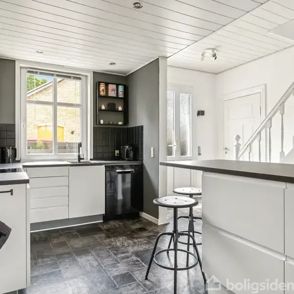 Et moderne køkken er indrettet med hvide skabe og sorte kakler, to krus med teksten "HOT" hænger ved ovnen, og en bar med to høje taburetter befinder sig i forgrunden.