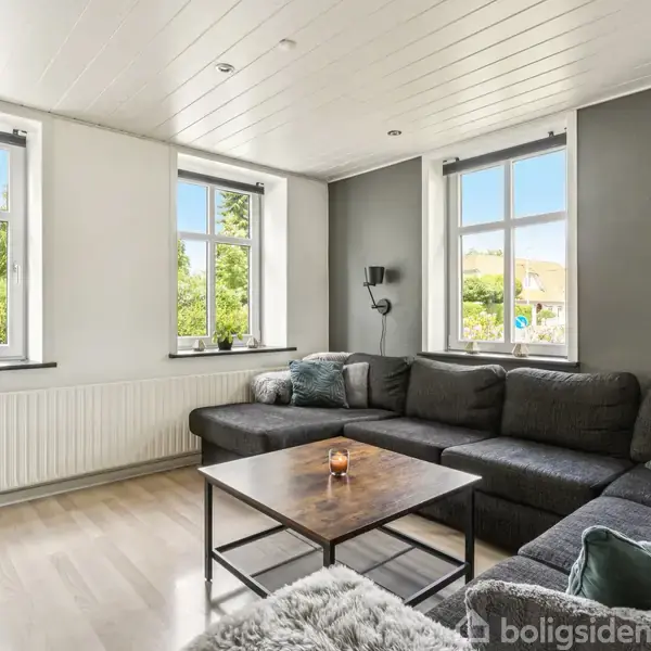 Stor hjørnesofa fyldt med puder står mod en lysegrå væg under tre vinduer. Et rektangulært sofabord foran sofaen har et tændt stearinlys. Fladskærms-tv til venstre.