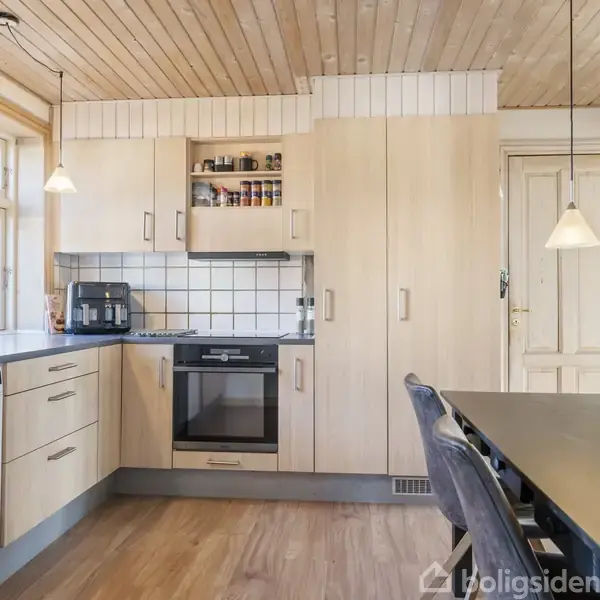 Køkken med lyse træskabe og -loft, opvaskemaskine, ovn, og mikrobølgeovn. Et rundt ur hænger på væggen. Bord med blå blomster i forgrunden.