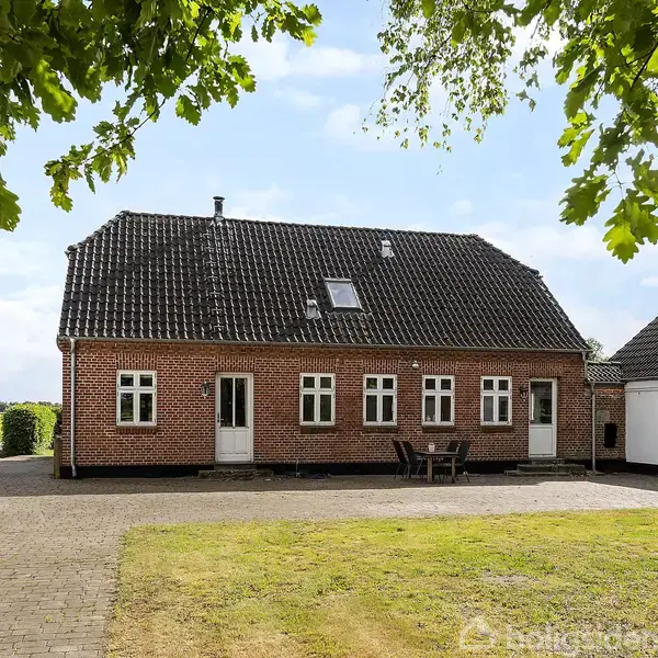 Brun murstenshus med skråt tag står stille på en gårdsplads med flisebelægning. Grønne marker og træer omkranser ejendommen, mens havemøbler ses foran huset.