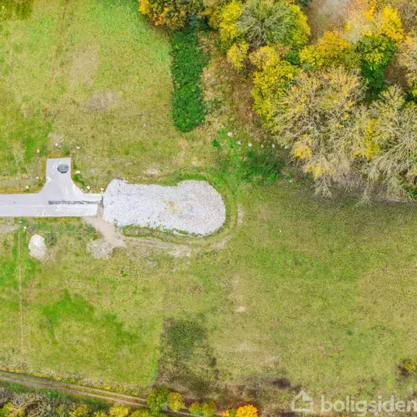 En grusbelagt indkørsel støder op til en stor græsmark omkranset af træer i efterårsfarver ved siden af et moderne hus med hvide vægge og sorte tage.