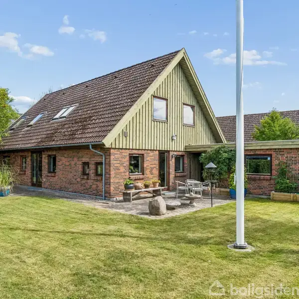 Et rødt murstenshus med spidst tag står i en grøn have med græsplæne og træer. Foran huset er der en terrasse med havemøbler.