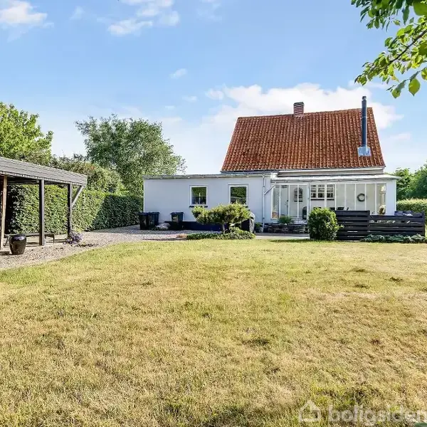Et hvidt hus med rødt tag står i en grøn have. En garagebygning er til venstre. Haven er omgivet af træer og buske under en blå himmel. Ingen tekst til stede.