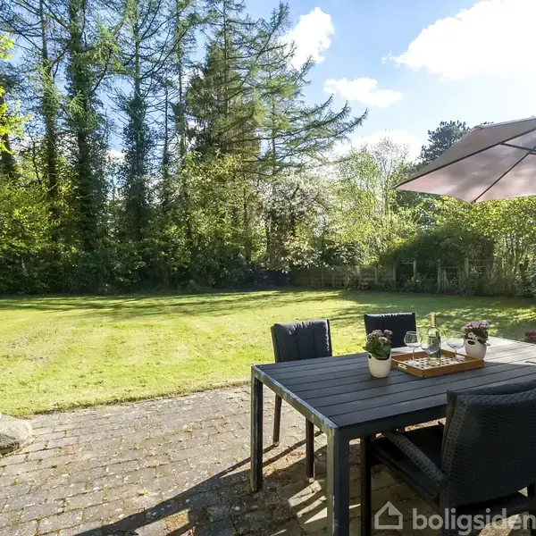 Terrassemøbler står på en flisebelagt patio med et bord, fire stole og en parasol. I haven omkring er der grøn græsplæne og høje træer i baggrunden under en blå himmel.