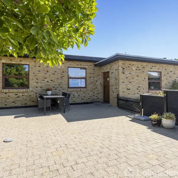 En gul murstensvilla står stille i solrig baggård med flisebelagt terrasse. Et sæt havemøbler er placeret foran huset, omgivet af grønne planter og en blå himmel i baggrunden.