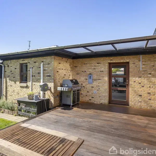 Terrasse med trædæk, omkranset af gule murstensvægge. En grill og plantebede står langs væggen. Til venstre ses en lille sort træbygning med rødt skilt.