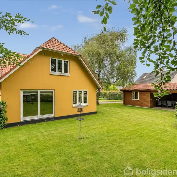 Gulmalet hus med saddeltag står i en velplejet have med græsplæne. Ved siden af huset ses en garage eller udhus. Himlen er blå med et par skyer.