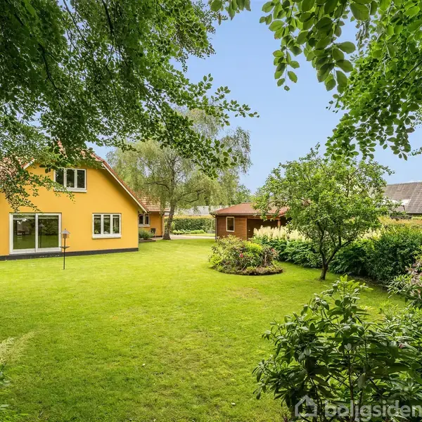 Huset er malet gult og står midt i en velplejet have med grøn græsplæne. Buske og træer omgiver haven, mens en mindre bygning ses i baggrunden.