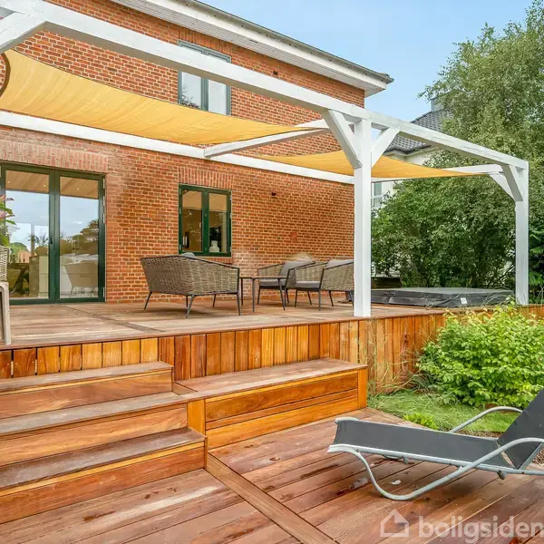 Terrasse med brune møbler under sejlskygger, placeret foran et murstenshus. Trætrapper fører til en liggestol på græsplænen, omgivet af grønne buske og træer.