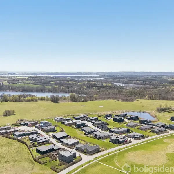 Boligområde set fra oven med huse, der står stille i en grøn, landlig kontekst med søer og skove i baggrunden under en klar blå himmel.