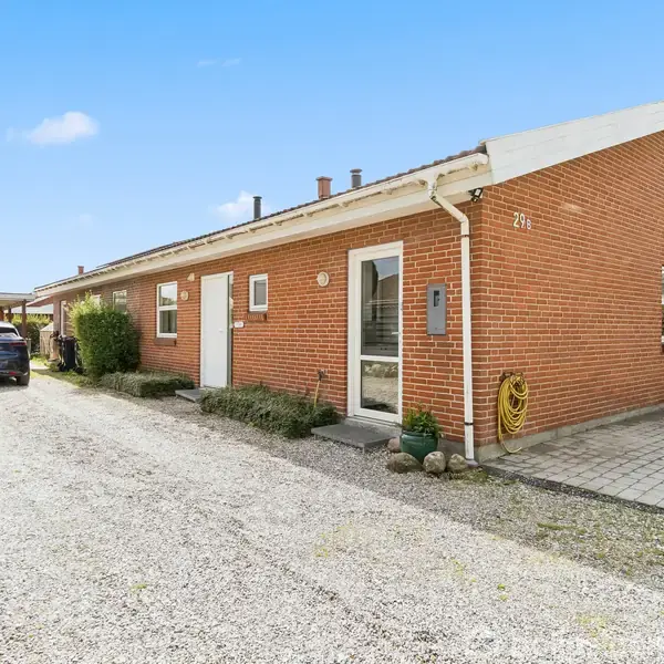 Rødt murstenshus står stille i en indkørsel, omgivet af grus og få buske. En sort bil er parkeret til venstre. Tallet "21" er synligt på facaden.