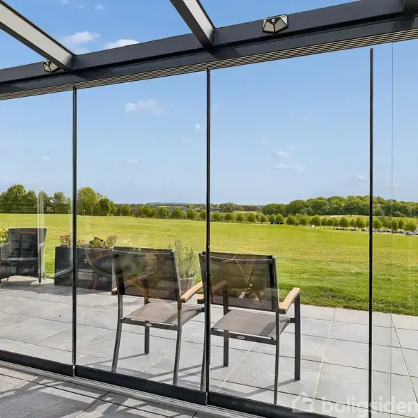 To stole står i en glasindhegnet terrasse med udsigt over en grøn græsplæne. Baggrunden viser en bred landskabsudsigt med træer og en blå himmel.