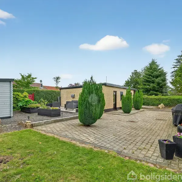 Flisebelagt terrasse med to små, kegleformede buske og et sort bord-sæt. Omgivet af en græsplæne og en gul murstensbygning samt en hvid garage i baggrunden.