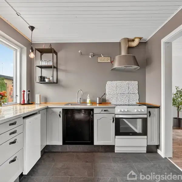 En moderne køkkenindretning med hvide skabe står ved en vindue, som lukker naturligt lys ind. Sort opvaskemaskine og komfur er indbygget. Køkkenets vægge er gråmalede.