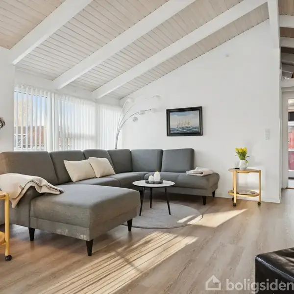 En grå hjørnesofa står i en stue med loft til kip og trælaminatgulv, flankeret af et rundt sofabord og små sideborde. Sollys strømmer ind gennem store vinduer.