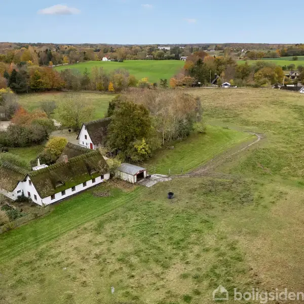 En traditionel gård med stråtag ligger blandt åbne marker og træer, omgivet af en grusvej i et landligt landskab.