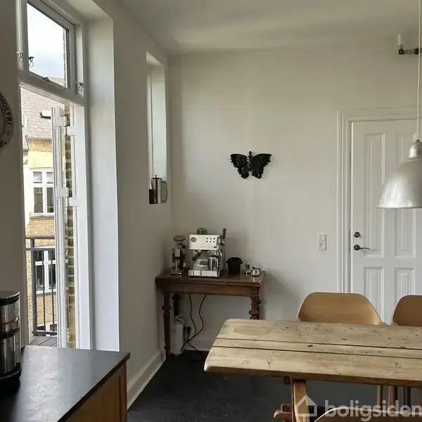 En kaffemaskine står på et lille træbord. På væggen hænger en sort sommerfugl dekoration. I rummet er der en åben dør til en balkon og spisebordsstole.