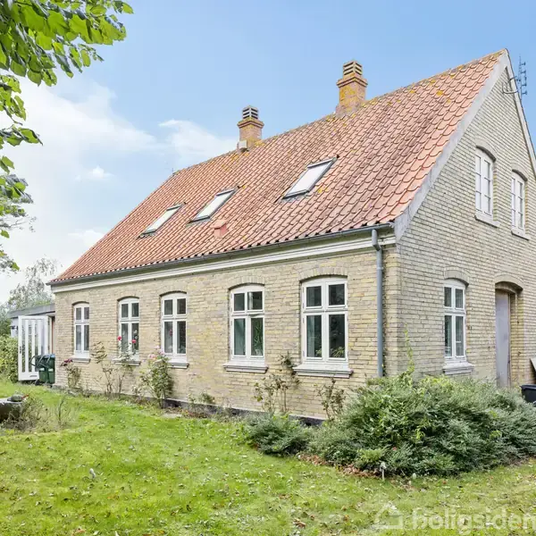 Et charmerende hus med rødt tegltag og mursten, har flere vinduer og en havedør. Det er omgivet af en grøn have med buske og træer under en klar, blå himmel.