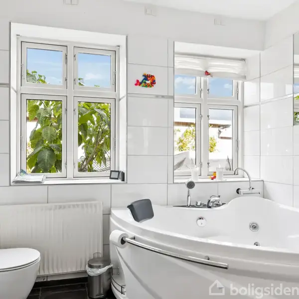 Boblebad står stille i et lyst badeværelse med hvide fliser. To vinduer bagved viser grønne blade udenfor. Toilettet og et skab er til venstre, spejl over boblebadet.