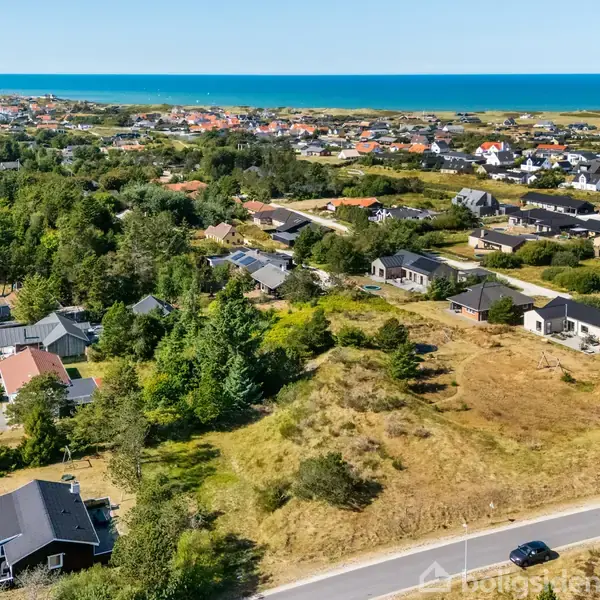 En landsby med huse spredt blandt træer og marker strækker sig mod kysten. Havet er synligt i baggrunden, mens en vej ses i forgrunden. Tekst ignoreret grundet retningslinjer.
