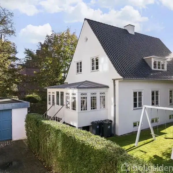 Hus med hvid facade og sort tegltag. Ved siden af er en blå garageport. En have med græsplæne og gyngestativ omgiver huset, og himlen er let skyet.