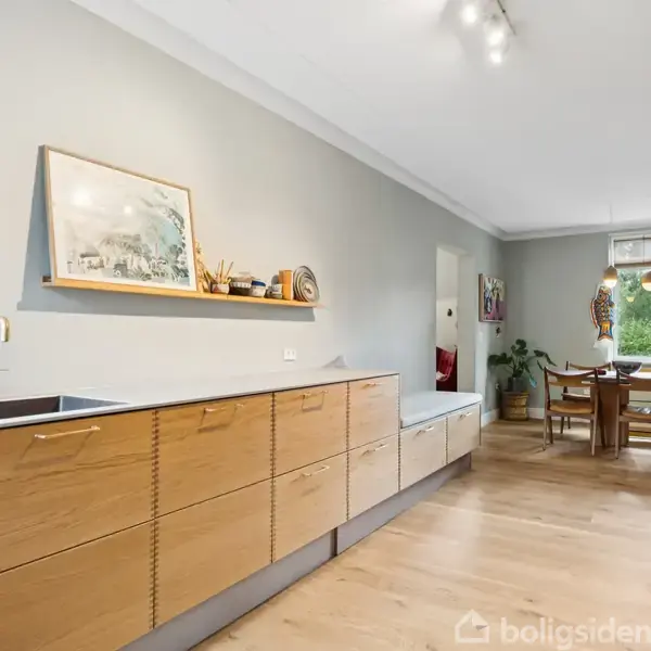 Køkken med træskabe langs væggen, håndvask til venstre og åbent mod spisestue med træbord og stole. Lysindfald fra vinduerne bagest i rummet.