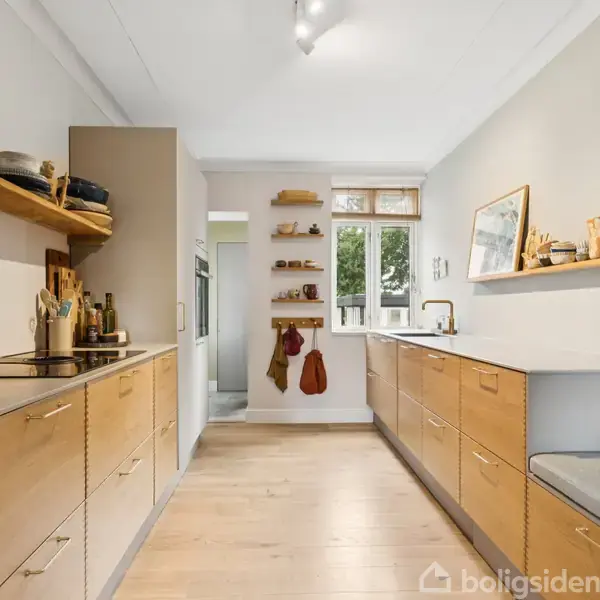 Køkkenet med træskuffer og åbne hylder rummer en kaffemaskine og køkkengrej. Store vinduer og lyse trægulve skaber en lys atmosfære. Tre mellemlange hylder bærer dekorationer.