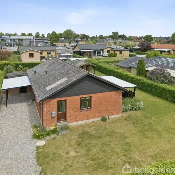 Rødt murstenshus står på en velholdt græsplæne, flankeret af grusindkørsel. Taget er mørkt med tagvinduer. Hustet omgives af lave hække og ligger i et villakvarter med andre boliger.