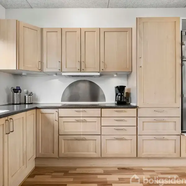 Et køkken med lyse træskabe står langs væggen. En kaffemaskine er placeret på bordpladen. Til venstre er et vindue, der giver naturligt lys.