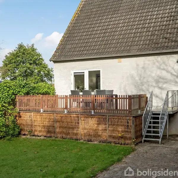 Hus med en træterrasse udstyret med stole og bord, på en velplejet græsplæne. En metaltrappe fører fra terrassen til haven. Baggrunden har træer og buske under en blå himmel.