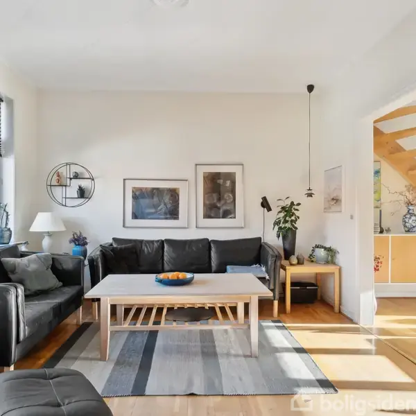 Sort lædersofa står stationært i en lys stue. Der er et træbord foran og malerier på væggen. Trappen fører til en overetage, ved siden af en plante.