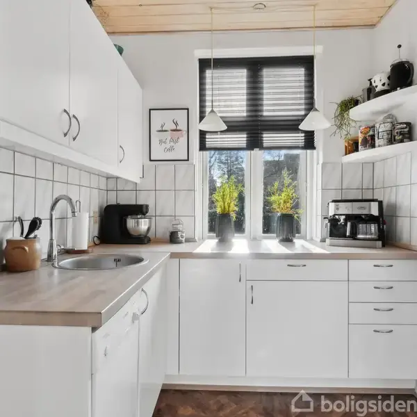 Et hvidt køkken med skabe og skuffer udfører funktionen af opbevaring. Bordpladen har en vask og køkkenmaskiner. En kaffemaskine står ved vinduet, der lader sollys strømme ind. Hylderne rummer krukker og dekorationer. teksten på plakaten: "But first, coffee".