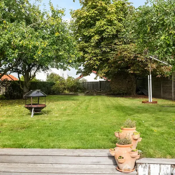 Græsplæne med et bålfad i midten, omgivet af frugttræer og hegn. Foran står en blomsterkrukke på en træterrasse. Klar, lys dag med delvis overskyet himmel.