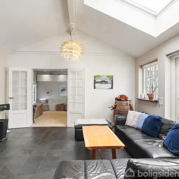 Sort læderhjørnesofa står i en stue med store vinduer og et fladt sofabord foran. Rummet har et lyst loft med en hængende lampe og åben dør til køkken.