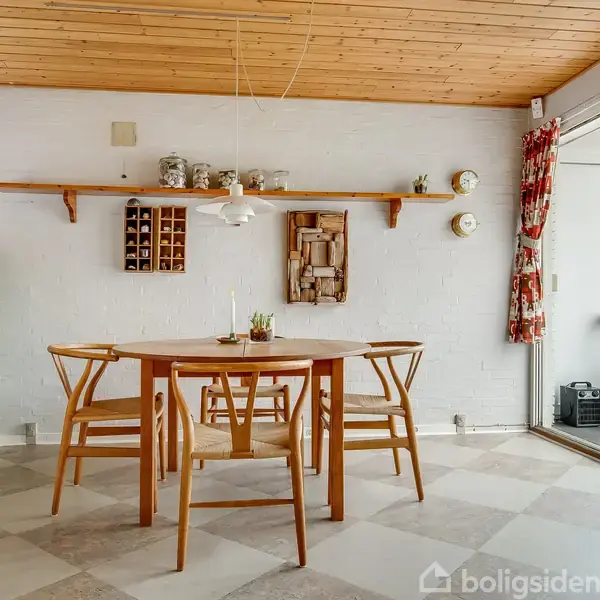 Et rundt træbord med stole står i et lyst rum med glasvægge og synlige træbjælker. Rummet er fyldt med grønne planter i krukker, hvilket skaber en naturinspireret atmosfære.