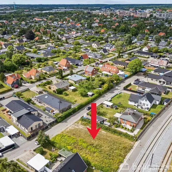 Tomt markeret med en rød pil; omgivet af huse i en forstad. Langs højre side løber en vej med spor. Grønt område og træer i baggrunden.
