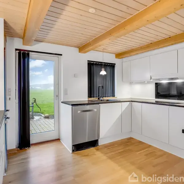 Et moderne køkken med hvide skabe, sort bordplade og en indbygget ovn. Køkkenet har en opvaskemaskine og åbner ud til en terrasse med udsigt til en grøn mark.