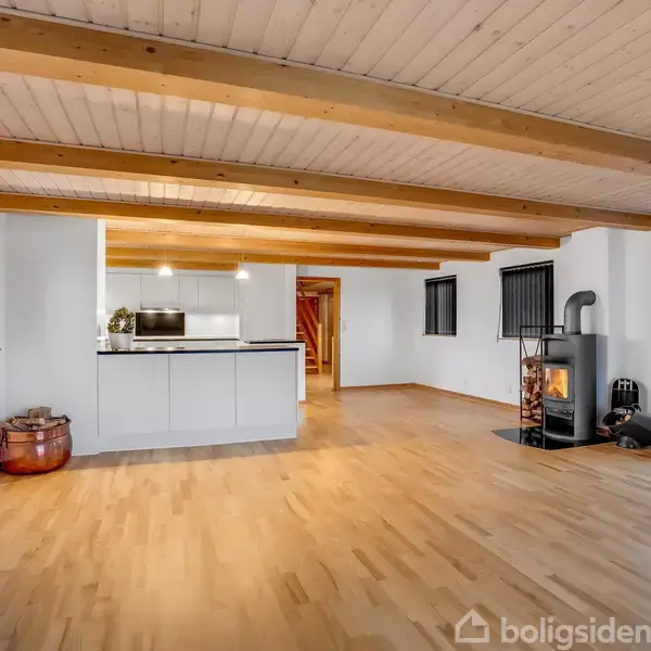 Brændeovn står tændt på lyst trægulv, opvarmer stue i moderne hus med hvide vægge, synlige bjælker i loftet, åbent køkken og små vinduer med skodder.