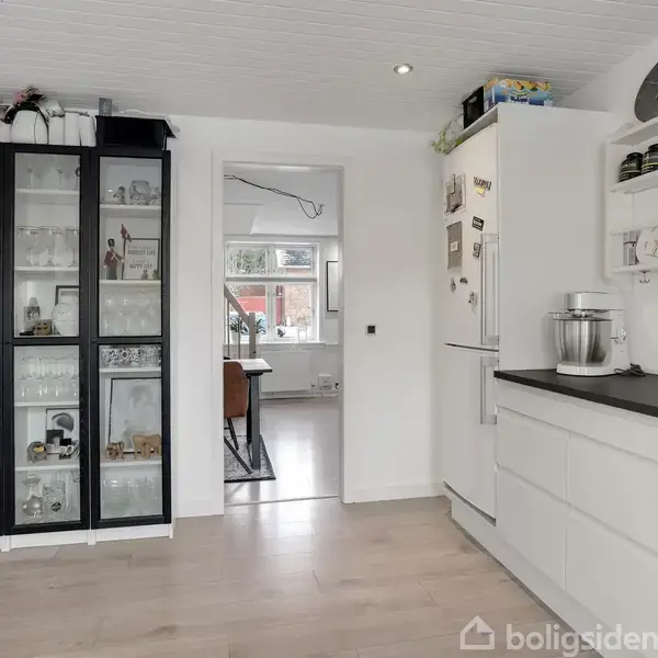 Hvidmalet, to-etagers hus med rødt tegltag i snefyldt indkørsel, omgivet af grønne buske. Nabohus er rødt mursten og også dækket af sne.