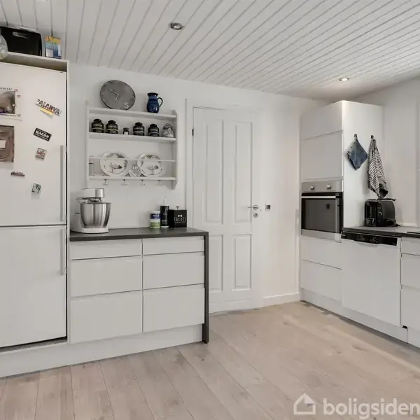 Et hvidt køkken med hvide skabe, en stående mikser og en ovn. Køleskabet er dekoreret med magneter. Rummet har lyst trægulv og hvid loftbeklædning.