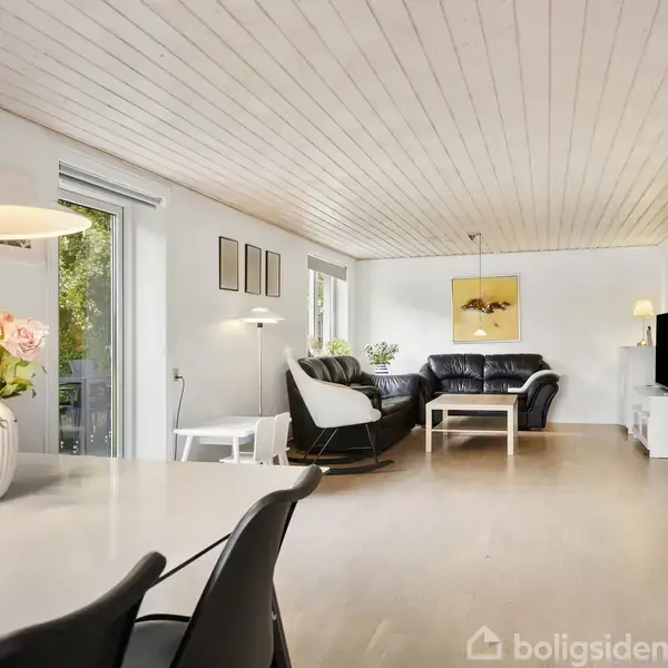 Blomster i vase står på et spisebord i et lyst opholdsrum med hvide vægge og træloft. Sofaer og sofa bord er arrangeret modsat tv og reoler.