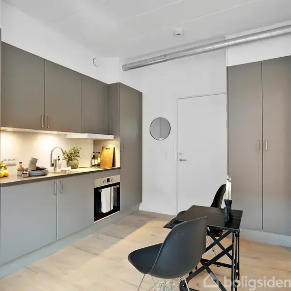 Et moderne køkken med grå skabe arrangeret langs en væg. En ovn og vask er integreret i skabene. To sorte stole står ved et lille bord i en lys lejlighed.