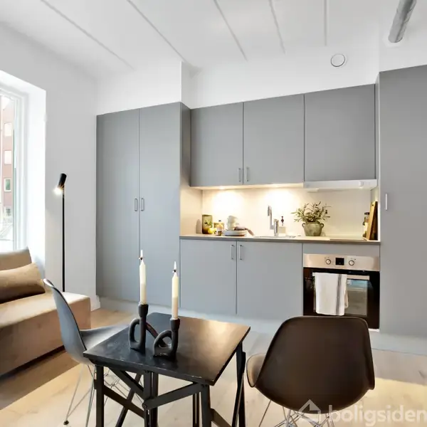 En lille stue-køkken med grå skabe og indbygget ovn. En grå sofa står ved vinduet til venstre. Der er et lille bord med to stole foran sofaen.