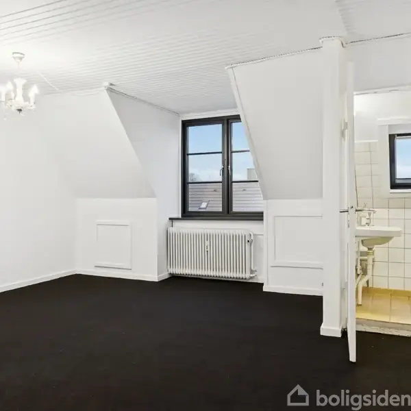 Et tomt værelse med sort gulvtæppe og hvidmalede vægge. Der er et stort vindue ved siden af en radiator. Til højre ses en åben dør til et badeværelse med fliser.