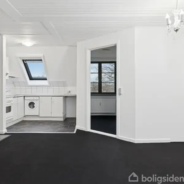 En stue med sort gulv og hvidmalede vægge. En dør fører til et køkken med vaskemaskine og ovenlys. Der er en lysekrone i loftet.